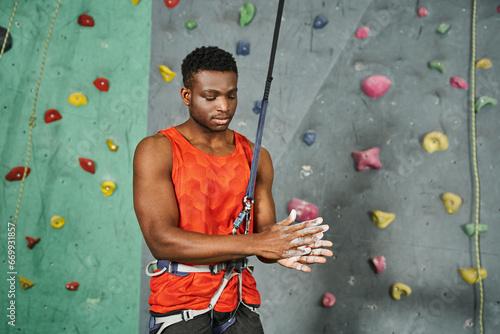 young athletic african american man in orange shirt with safety rope using talc powder, bouldering