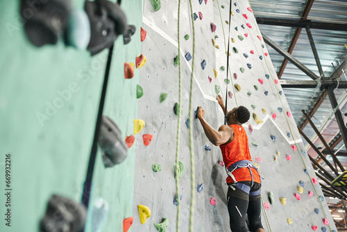 strong young african american man in orange shirt with alpine harness ascending up rock wall