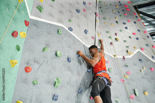 sporty young african american man using safety rope and harness to climb up rock wall, bouldering