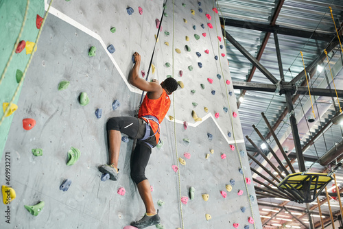 strong african american man in orange shirt using safety rope and alpine harness to climb up wall