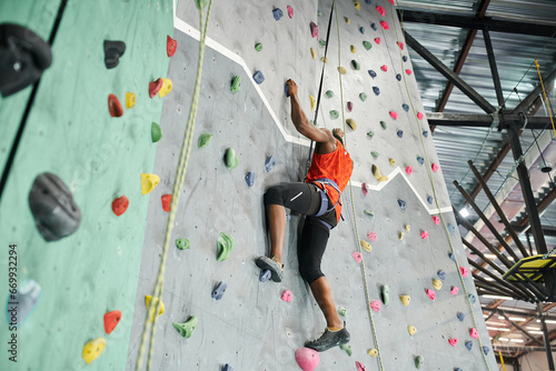 strong athletic african american man with alpine harness climbing up rock wall gripping on rocks