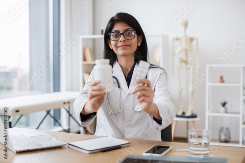 Friendly female therapist in glasses showing white jar of medicine while sitting at desk. Experienced indian woman in white coat recommending best alternative of treating at doctor's office.