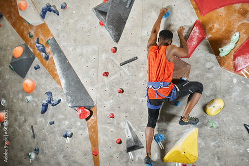 young african american man with alpine harness ascending up climbing wall, back view, bouldering