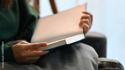Carefree young woman in warm sweater reading book on comfortable couch at home