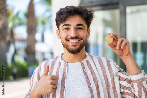 Handsome Arab man holding a Bitcoin at outdoors with thumbs up because something good has happened