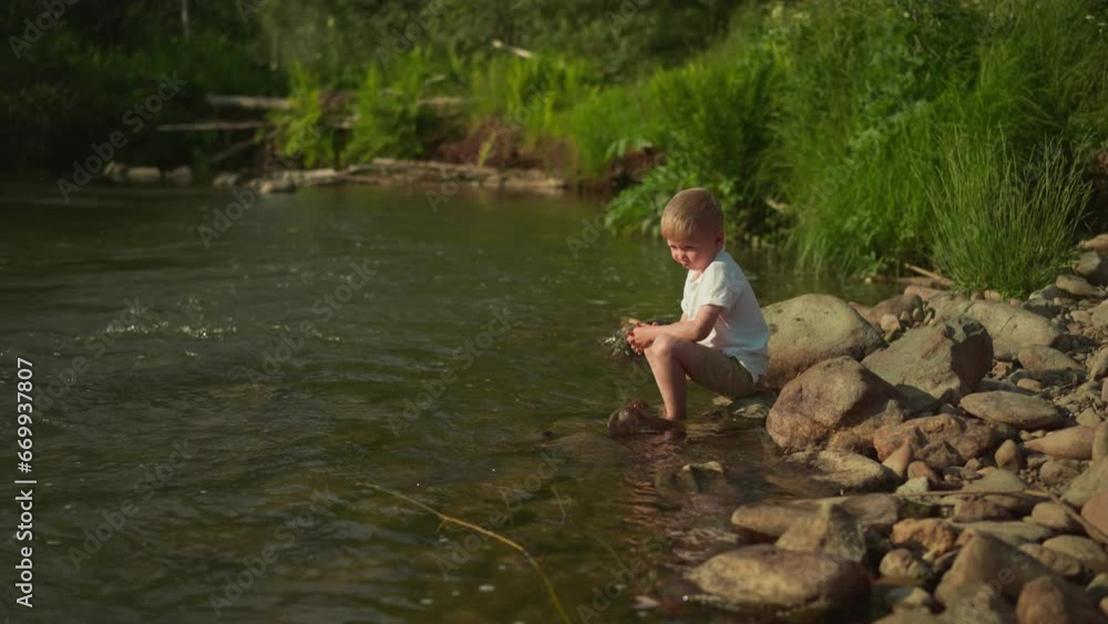 Little boy plays with pebbles resting on river bank on sunny day. Thoughtful child sits on big rock thinking about summer vacation. Concept of recreation and childhood Little boy plays with pebbles resting on river bank on sunny day. Thoughtful child sits on big rock thinking about summer vacation. Concept of recreation and childhood