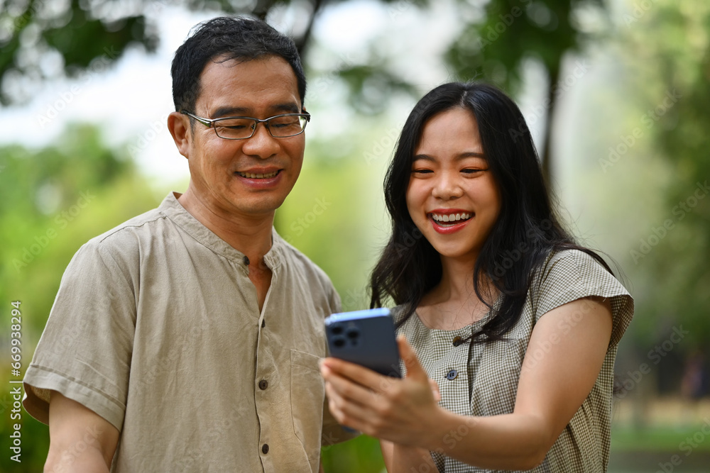 Cheerful young woman and middle age father watching online video in mobile phone while standing in the park Cheerful young woman and middle age father watching online video in mobile phone while standing in the park