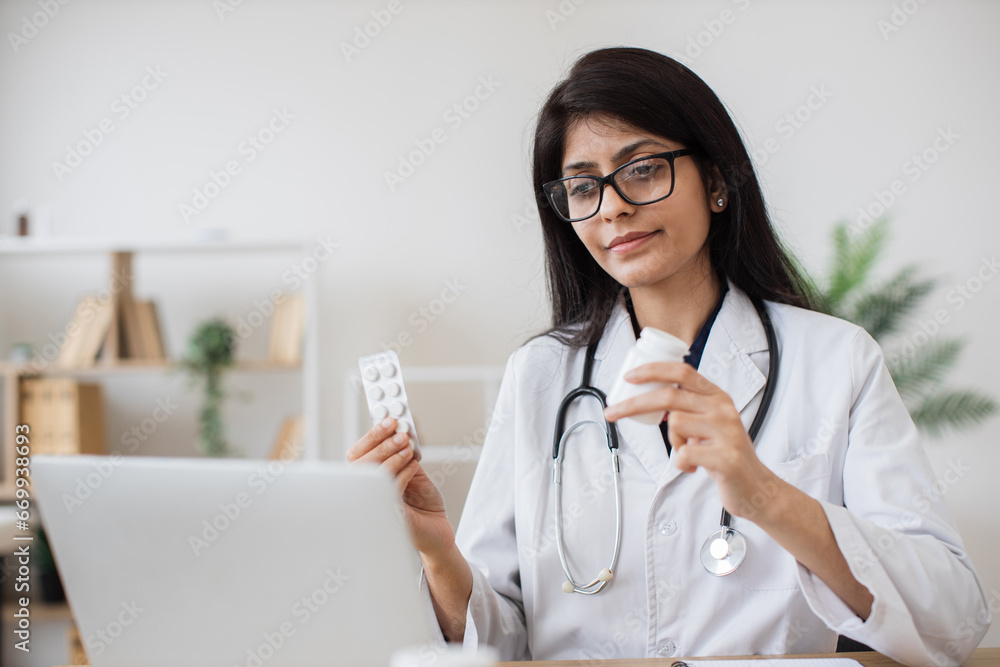 Hindu hospital worker talking to patient on video call and making consultation about treatment in bright office. Attentive nurse holding pills in hands and prescribing course of medication. Hindu hospital worker talking to patient on video call and making consultation about treatment in bright office. Attentive nurse holding pills in hands and prescribing course of medication.