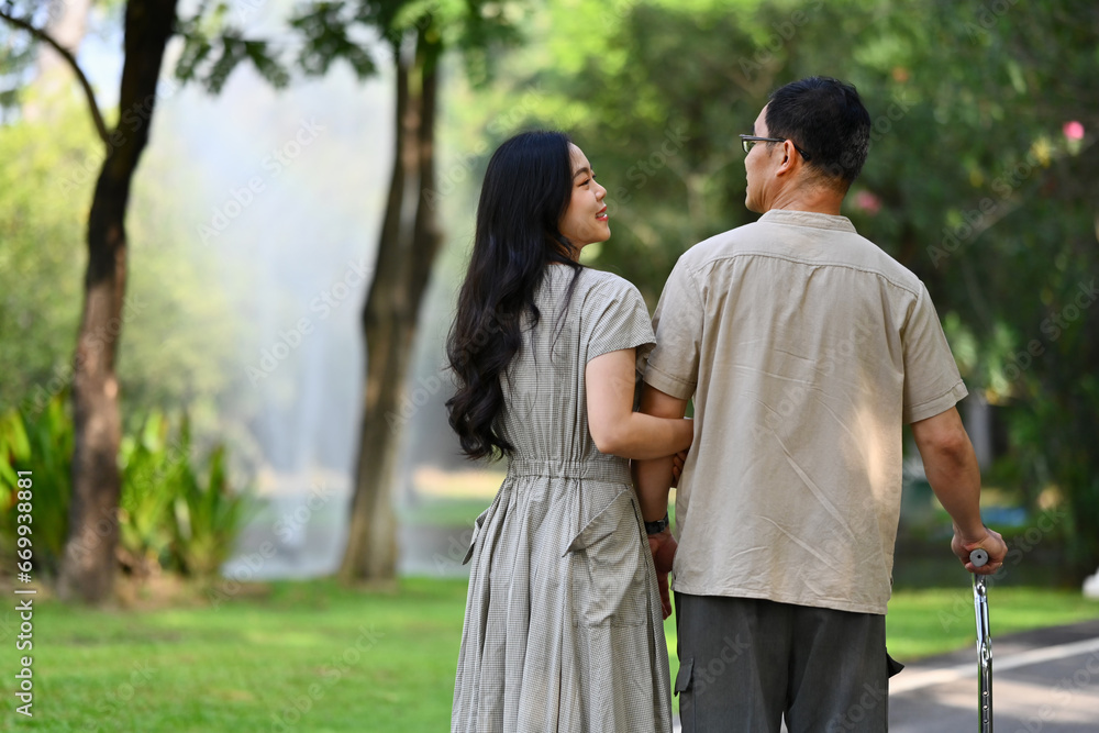 Rear view of caring daughter talking and walking with middle age father at park. Family, generation and authentic lifestyle Rear view of caring daughter talking and walking with middle age father at park. Family, generation and authentic lifestyle