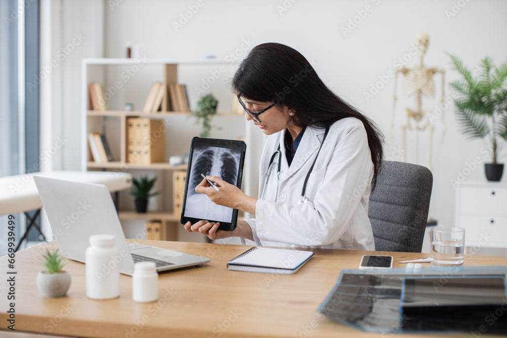 Concentrated female doctor talking on video call while showing result of examination of patient with tuberculosis. Indian lady consulting person using laptop and tablet in spacious clinic room. Concentrated female doctor talking on video call while showing result of examination of patient with tuberculosis. Indian lady consulting person using laptop and tablet in spacious clinic room.