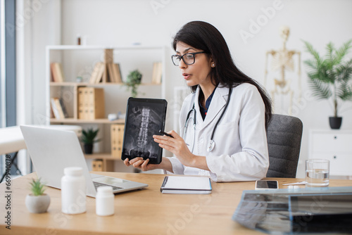 Competent female doctor discussing results of x-ray examination with patient on wireless laptop in cabinet. Attentive hindu woman holding tablet with image of spine and providing consultation.