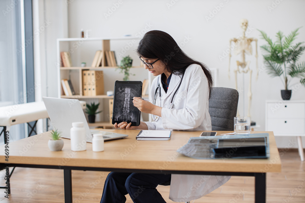 Competent female doctor discussing results of x-ray examination with patient on wireless laptop in cabinet. Attentive hindu woman holding tablet with image of spine and providing consultation. Competent female doctor discussing results of x-ray examination with patient on wireless laptop in cabinet. Attentive hindu woman holding tablet with image of spine and providing consultation.