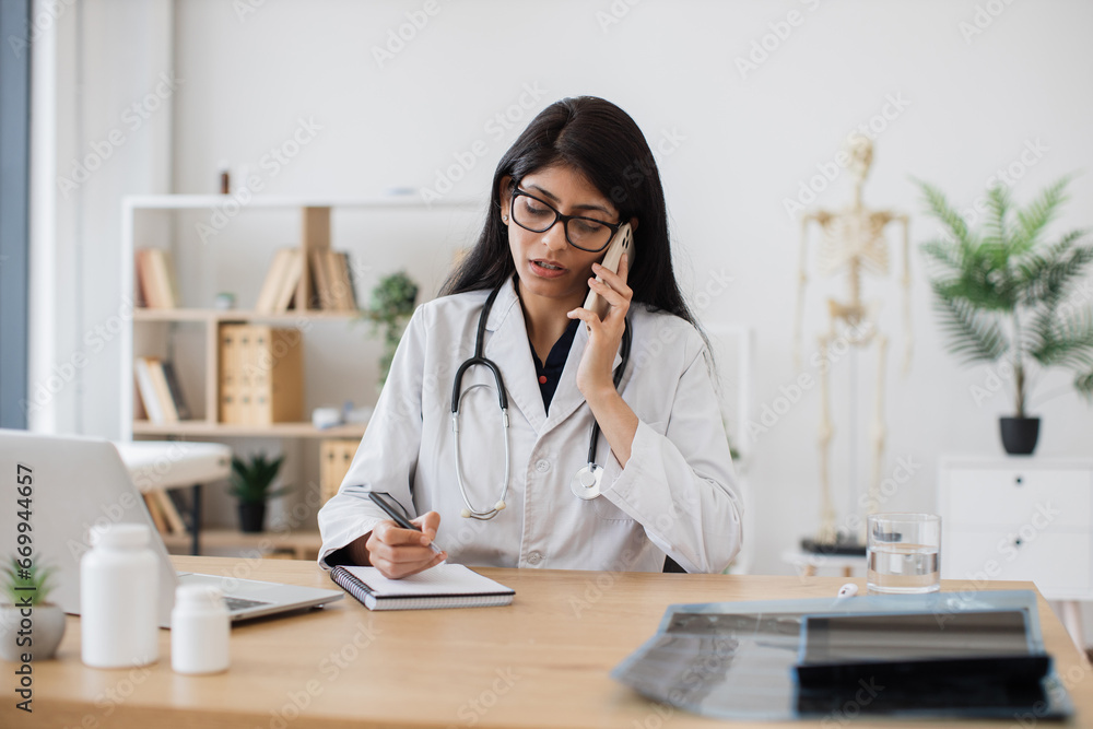 Experienced indian doctor talking on modern smartphone with ill patient while sitting at personal workplace in modern hospital. Mature female writing data about sick person in own notebook indoors. Experienced indian doctor talking on modern smartphone with ill patient while sitting at personal workplace in modern hospital. Mature female writing data about sick person in own notebook indoors.