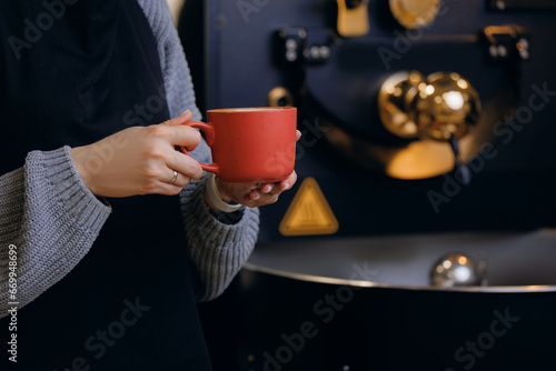 Concept production and roasting craft coffee in small workshop. Worker holds cup in front of roaster machine
