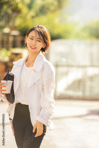 햇빛이 비치는 공원에서 검정 가방을 겨드랑이에 끼고 한손으로 커피를 든 채 밝게 웃고 있는 젊은 한국 여성