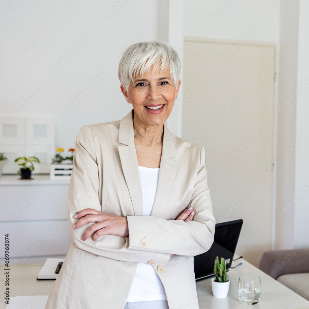 © Jelena Stanojkovic - Confident female looking at camera while standing in home office. Cropped portrait of an attractive senior businesswoman sitting at the desk with her arms crossed while working from home.