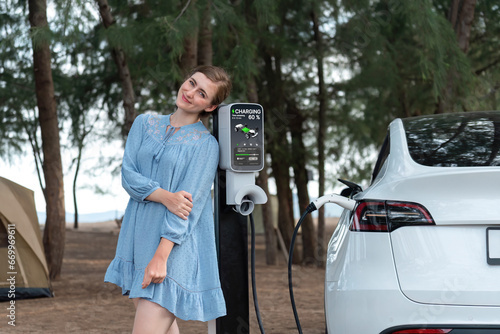 Holiday road trip vacation traveling to the beach camp with electric car, young woman recharge EV vehicle with green and clean energy. Beach travel camping with eco-friendly EV car .Perpetual