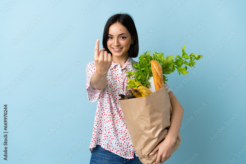 Young woman holding a grocery shopping bag doing coming gesture Young woman holding a grocery shopping bag doing coming gesture
