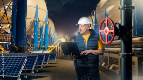 Man is industrialist. Guy with laptop on territory of plant. Factory engineer with computer. Energy manufactory in evening. Industrialist man walks around factory grounds. BPVC tanks. Solar panels photo