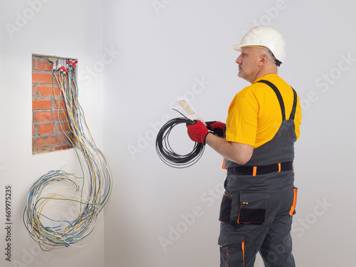 Man is electrician. Construction worker with power wire. Electrician near power panel area. Worker prepares to install electrical equipment. Man electrician in hardhat and gray overalls