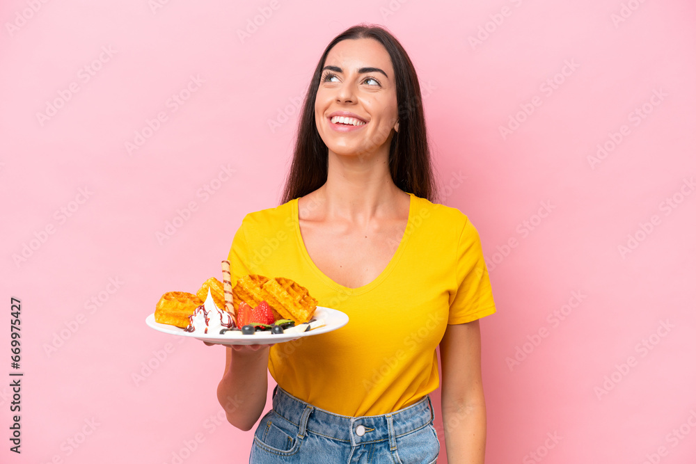Young caucasian woman holding waffles isolated on pink background thinking an idea while looking up Young caucasian woman holding waffles isolated on pink background thinking an idea while looking up