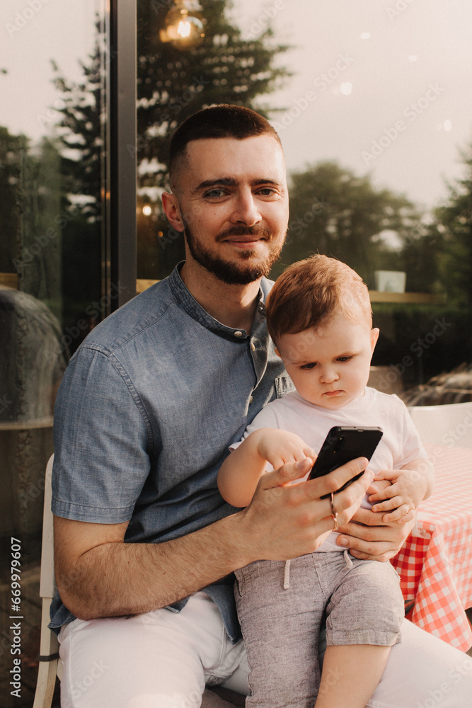 Fototapeta premium Young man using a phone mobile for work and holding his son on his hand on the cafe. Being a daddy and working everywhere. Freelancing and networking concept