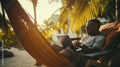 A young man with a laptop works on the ocean shore. Freelancer man relaxing at the resort. Remote work concept, vacation. © Alina Tymofieieva