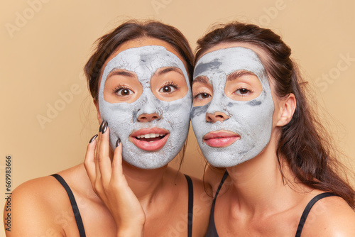 Two cute girls make cosmetic masks on their faces, look at the camera with interest, each has mask of medicinal clay on her face, skincare products concept, copy space