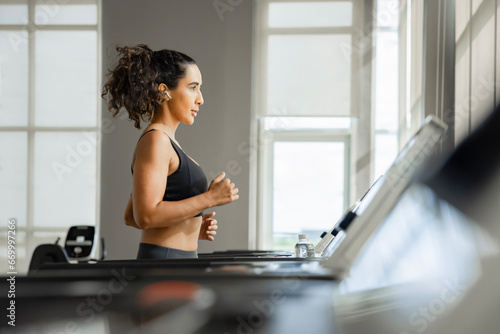 Muscular fitness woman listening to music on wireless earphones while running on a treadmill, doing exercises in the morning at a gym. 