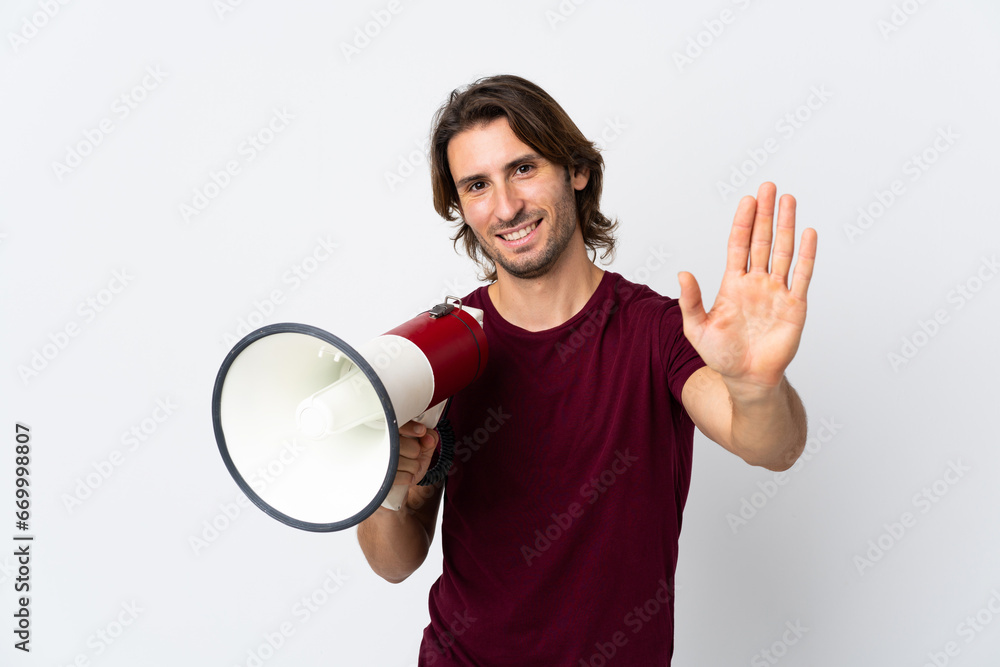 Young handsome man isolated on white background holding a megaphone and saluting with hand with happy expression Young handsome man isolated on white background holding a megaphone and saluting with hand with happy expression