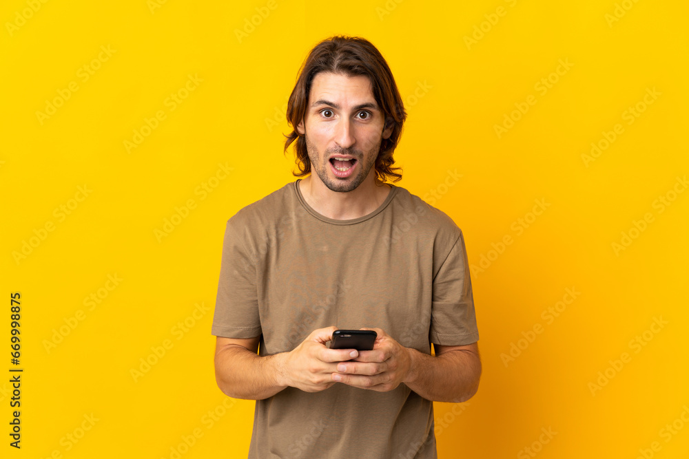 Young handsome man isolated on yellow background surprised and sending a message Young handsome man isolated on yellow background surprised and sending a message