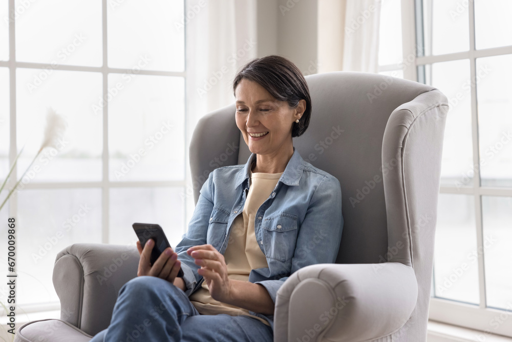 Smiling middle-aged female sit rest on cozy armchair with modern cellphone, enjoy chat with grown up children or friends, share messages use messenger, calling or make order, buy services on internet Smiling middle-aged female sit rest on cozy armchair with modern cellphone, enjoy chat with grown up children or friends, share messages use messenger, calling or make order, buy services on internet