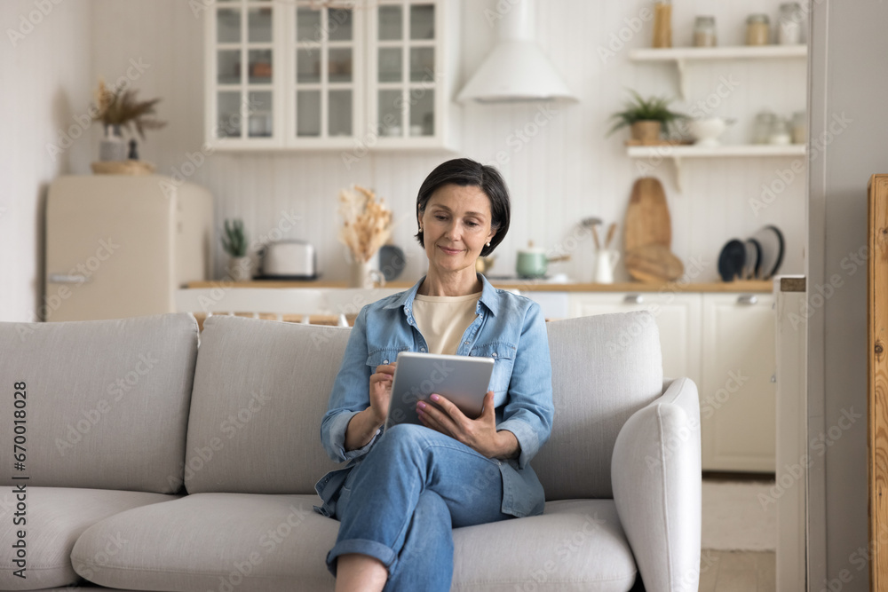 Pretty middle-aged woman spending leisure on internet sit on couch using digital tablet, choose goods, buy e-services through e-commerce app or website, enjoy easy remote electronic shopping at home