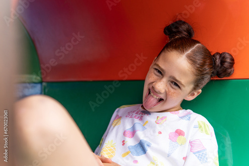 Portrait of a crazy red haired girl with freckles laughing and sticking her tongue out inside a tube in the playground, concept of happiness and joy