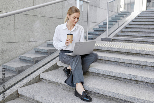 Confident blond young business woman sitting on stairs in the city and checking laptop
