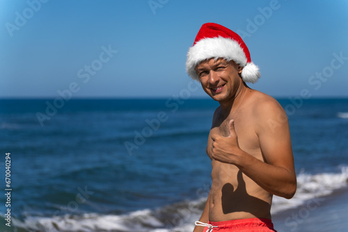 Young man in coral-colored swimming shorts and a Santa Claus hat shows a thumbs up on the seashore. Christmas holidays