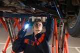 A young female car mechanic with a flashlight inspects the bottom of the car that is on the lift. Suspension and brake system diagnostics
