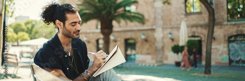 Young italian guy with ponytail and stubble sits on street bench and makes sketches with pen on piece of paper