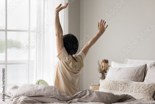 Rear view mature woman in comfort pajama awakening after healthy night enough sleep in the morning in bedroom, waking up, feels refreshed, stretching her body seated in bed, starting new day at home