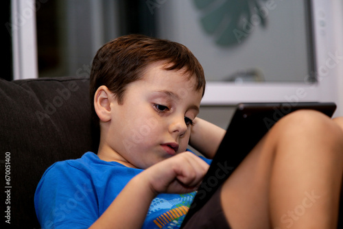 A boy sitting on a sofa at home playing with a tablet. Serious expression on his face. Concept of addiction to electronic games in young children.