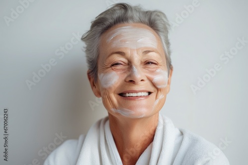 A beautiful elderly gray-haired woman with a cosmetic nourishing moisturizing mask on her face smiles on gray background. Facial skin care concept, beauty, care, cosmetology