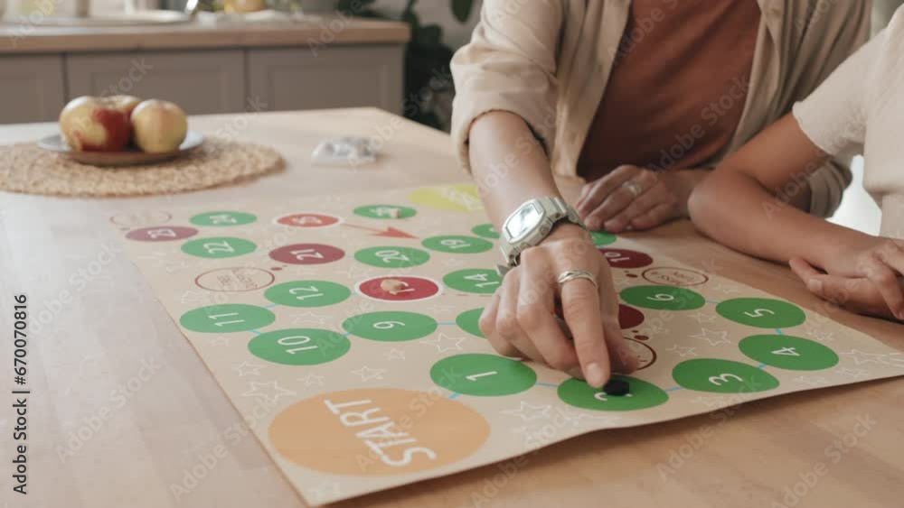 Slowmo of unrecognizable young woman and her daughter sitting at wooden table in kitchen playing board game with dice, chips and colorful sheet of paper with numbers in circles Slowmo of unrecognizable young woman and her daughter sitting at wooden table in kitchen playing board game with dice, chips and colorful sheet of paper with numbers in circles