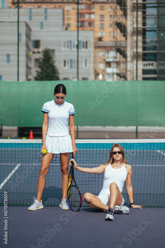 Photo of two beautiful female friends on the tennis court with rackets resting after training.