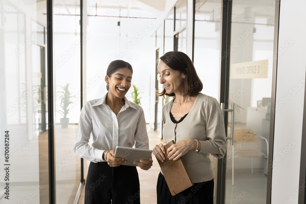Two positive diverse coworker women walking in office hall, using tablet computer, discussing online media content. Younger employee showing gadget screen to elder colleague, asking for advice Two positive diverse coworker women walking in office hall, using tablet computer, discussing online media content. Younger employee showing gadget screen to elder colleague, asking for advice