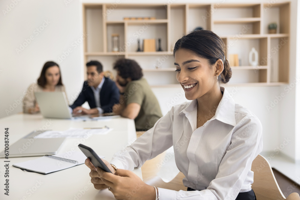 Happy confident business professional girl using smartphone over large office co-working table, sitting in meeting room with diverse colleagues talking in background, typing on cell, smiling Happy confident business professional girl using smartphone over large office co-working table, sitting in meeting room with diverse colleagues talking in background, typing on cell, smiling