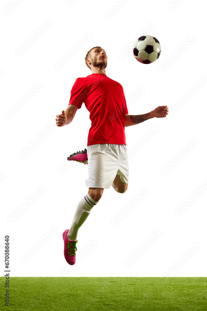 Young man, professional player in red sportwear and boots training football tricks against white background with green grass.