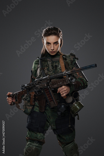 Portrait of a female soldier in military attire, holding a homemade automatic rifle, depicting a rebel or partisan in a Middle Eastern conflict against a gray background © Fxquadro