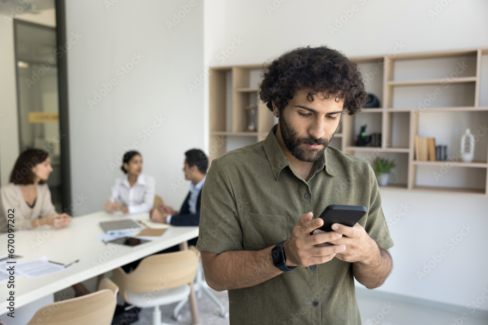 Focused young Arab business professional man typing on mobile phone while team on coworkers talking in background at large meeting table. Entrepreneur guy using online app on cellphone