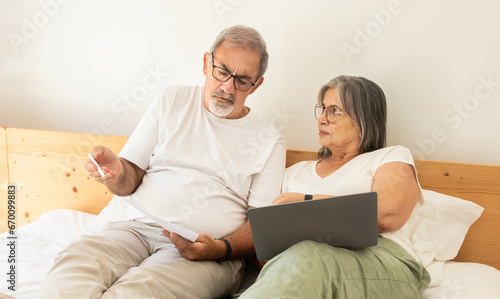 Sad confused old caucasian husband and wife lie on bed, pay for bills, taxes