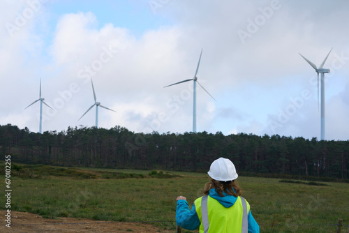 Rear view of female worker with a white helmet and yellow vest in front of wind turbines in a rural renewable energy area